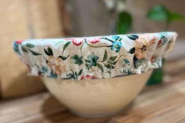Bowl with bread bowl cover on it. Cutting boards and flowers in the background.
