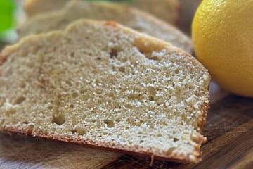 slices of pound cake on a wooden cutting board with a lemon on the side and plant leaves in the background