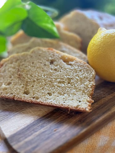 Slice of lemon pound cake on a cutting board with a lemon in the background and a leaf in the background