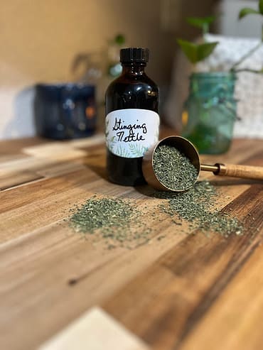 bottle on a counter with a copper measuring cup with loose herbs beside it - bottle is labeled "stinging nettle"