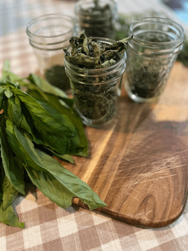 Several jars of dried herbs on a wooden cutting board with fresh basil to the side