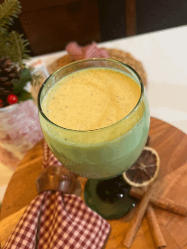 Eggnog in a glass on a wooden stand with cinnamon sticks and dried orange slices on the stand